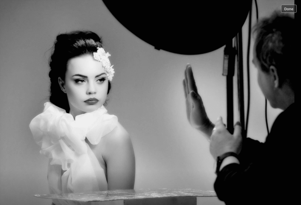 Behind-the-scenes photo of Peter Trainer directing a model during a studio beauty shoot with soft lighting and white styling elements.